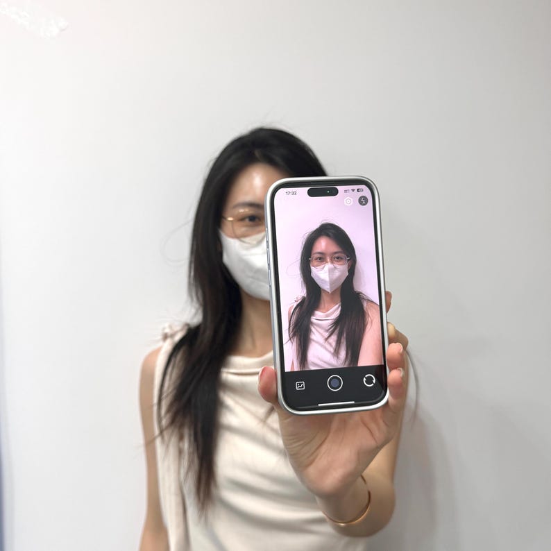 May include: A silver smartphone displaying a selfie of a person wearing glasses and a face mask. The phone is held up in front of a person wearing a mask and glasses. The background is a plain white wall.