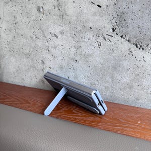May include: A gray Samsung foldable phone is propped up by a light blue stand. The phone is open and resting on a wooden surface, with a gray concrete wall in the background. The Samsung logo is visible.