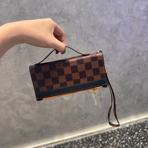 Peut inclure: Un portefeuille à carreaux marron et noir avec une bordure noire et un intérieur orange. Le portefeuille a une petite poignée et une dragonne. Le portefeuille est tenu par une personne.