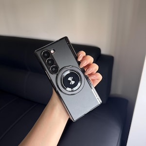 May include: A dark gray smartphone case with a silver-colored trim and a circular magnetic attachment. The case has a triple-lens camera design and a metallic ring with a decorative pattern. The phone case is held in a hand.