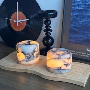 May include: Two cylindrical stone lamps with a white and dark gray marbled pattern emit a warm internal glow. These lamps are positioned on a light-colored, wavy-edged wooden board, accompanied by a black candlestick and a vinyl record clock.