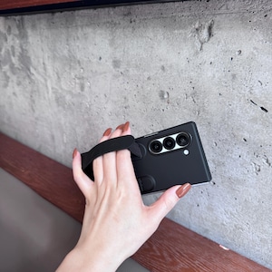 May include: Black smartphone case with a built-in black hand strap. The case has three camera lenses and a small white dot. The phone case is held against a gray concrete wall.