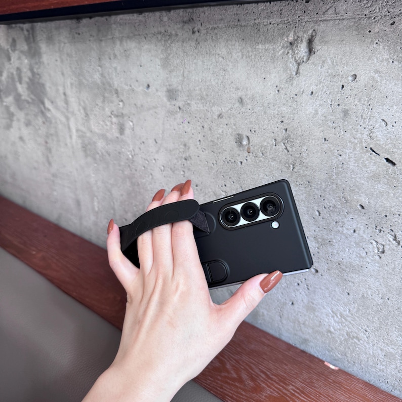 May include: Black smartphone case with a built-in black hand strap. The case has three camera lenses and a small white dot. The phone case is held against a gray concrete wall.