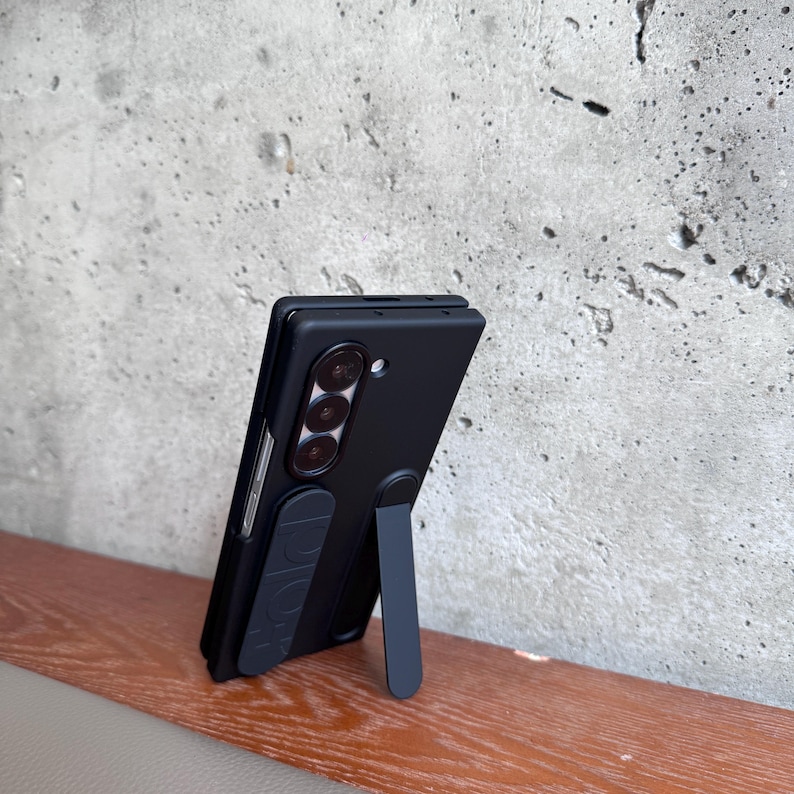 May include: Black phone case with a stand, featuring a triple-lens camera design. The case is propped up on a wooden surface against a gray concrete wall. The stand is extended, providing support for the phone.