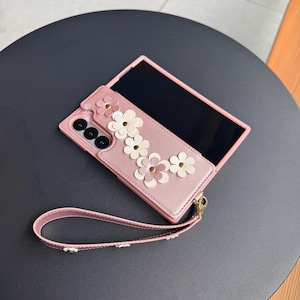 May include: Pink phone case with white and pink flower embellishments. The case has a matching wrist strap and is shown with a black screen. The case is on a dark gray surface.
