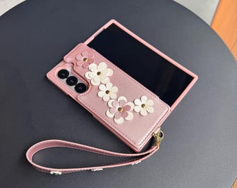 May include: Pink phone case with white and pink flower embellishments. The case has a matching wrist strap and is shown with a black screen. The case is on a dark gray surface.