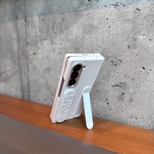 May include: A white phone case with a built-in stand, designed for a foldable phone. The case has a sleek, modern design with the word "FOLD" embossed on the side. The case is propped up on a wooden surface against a gray concrete wall.