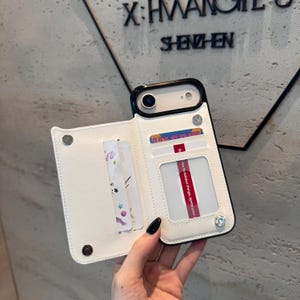 May include: A black and white phone case with a wallet design. The case has a white leather-like exterior with card slots and a clear ID window. The phone case is open, revealing the phone and cards.