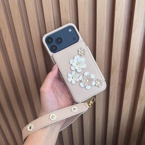 May include: A blush pink phone case with a floral design. The case features several 3D flowers in white, blush, and gold. A matching wrist strap with flower accents is attached. The phone case is held in a hand.