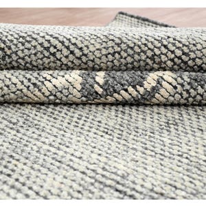 Puede incluir: Primer plano de una alfombra enrollada con un patrón texturizado. La alfombra presenta una combinación de gris claro, gris oscuro y blanco roto. El diseño parece ser un patrón repetitivo de formas pequeñas e irregulares.
