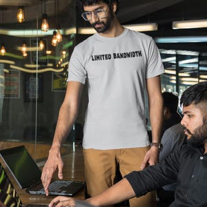 May include: Two people are working on laptops in an office setting. One person is wearing a grey t-shirt with the text "Limited Bandwidth" printed on it. The other person is wearing a black button-down shirt.