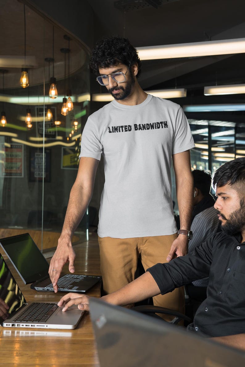 May include: Two people are working on laptops in an office setting. One person is wearing a grey t-shirt with the text "Limited Bandwidth" printed on it. The other person is wearing a black button-down shirt.