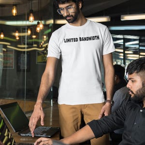 May include: Two people are working at a desk with laptops. One person is wearing a white t-shirt with the text "Limited Bandwidth" printed on it. The other person is wearing a black button-down shirt.