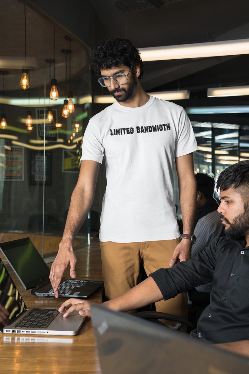 May include: Two people are working at a desk with laptops. One person is wearing a white t-shirt with the text "Limited Bandwidth" printed on it. The other person is wearing a black button-down shirt.