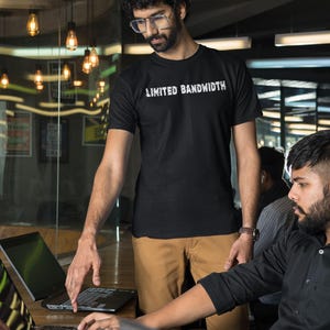 Pode incluir: Duas pessoas est&atilde;o trabalhando em laptops em um ambiente de escrit&oacute;rio. Uma pessoa est&aacute; vestindo uma camiseta preta com o texto "Limited Bandwidth" impresso nela. A outra pessoa est&aacute; vestindo uma camisa preta de bot&otilde;es.