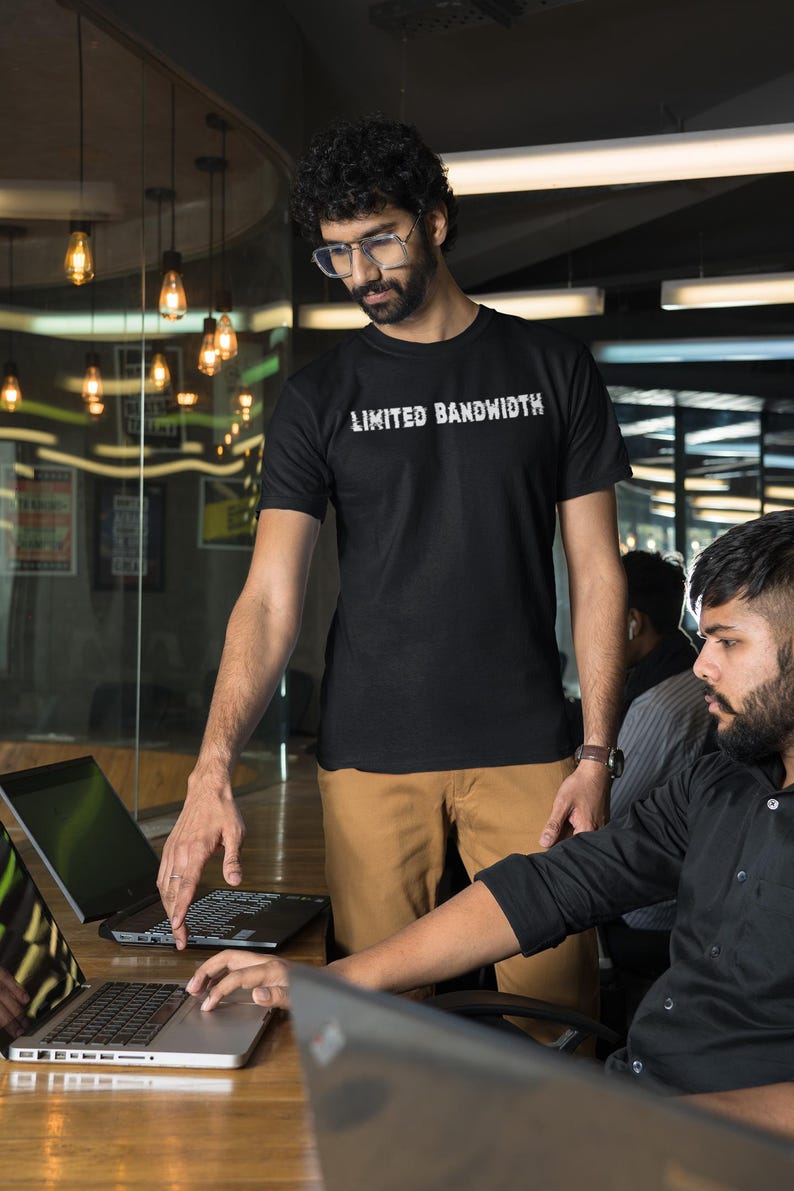 Pode incluir: Duas pessoas est&atilde;o trabalhando em laptops em um ambiente de escrit&oacute;rio. Uma pessoa est&aacute; vestindo uma camiseta preta com o texto "Limited Bandwidth" impresso nela. A outra pessoa est&aacute; vestindo uma camisa preta de bot&otilde;es.