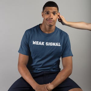 May include: A man wearing a navy blue t-shirt with the text "WEAK SIGNAL" in white. He is sitting on a wooden stool and has a confused expression on his face.