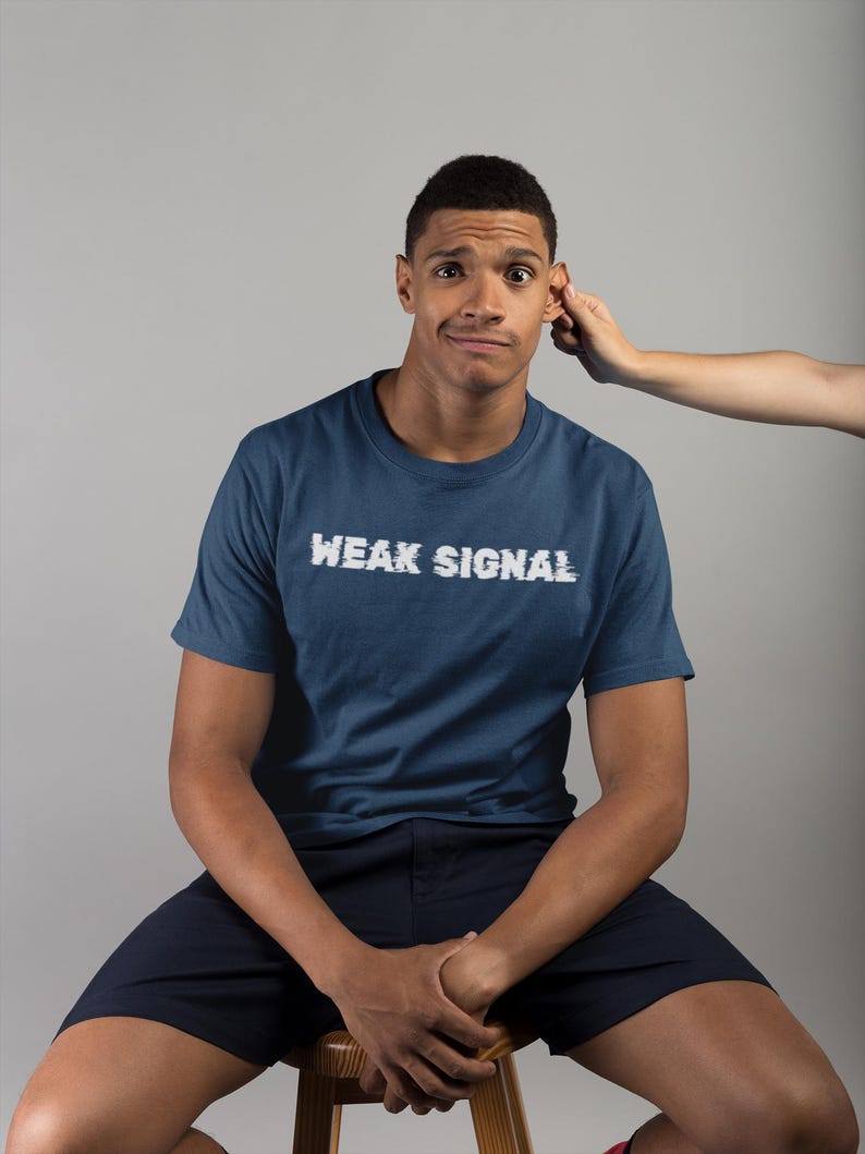 May include: A man wearing a navy blue t-shirt with the text "WEAK SIGNAL" in white. He is sitting on a wooden stool and has a confused expression on his face.