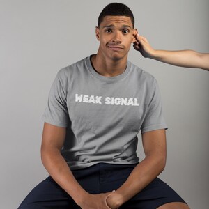 May include: A man wearing a grey t-shirt with the text "WEAK SIGNAL" in white lettering. He is sitting on a wooden stool wearing dark blue shorts.
