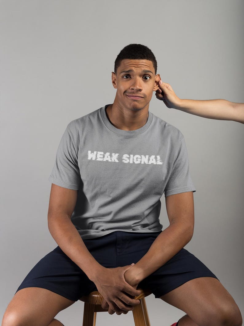 May include: A man wearing a grey t-shirt with the text "WEAK SIGNAL" in white lettering. He is sitting on a wooden stool wearing dark blue shorts.
