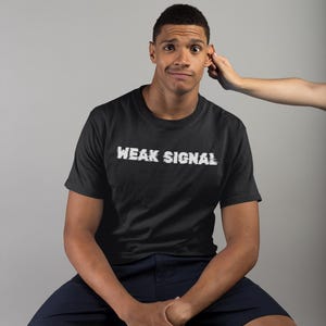 May include: A black t-shirt with the text "WEAK SIGNAL" in white lettering. The person wearing the shirt is sitting on a wooden stool.