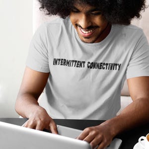 Puede incluir: Una persona que lleva una camiseta blanca con el texto "Intermittent Connectivity" impreso en ella, est&aacute; usando una computadora port&aacute;til. La persona est&aacute; sentada en una mesa con una taza de caf&eacute; sobre la mesa.