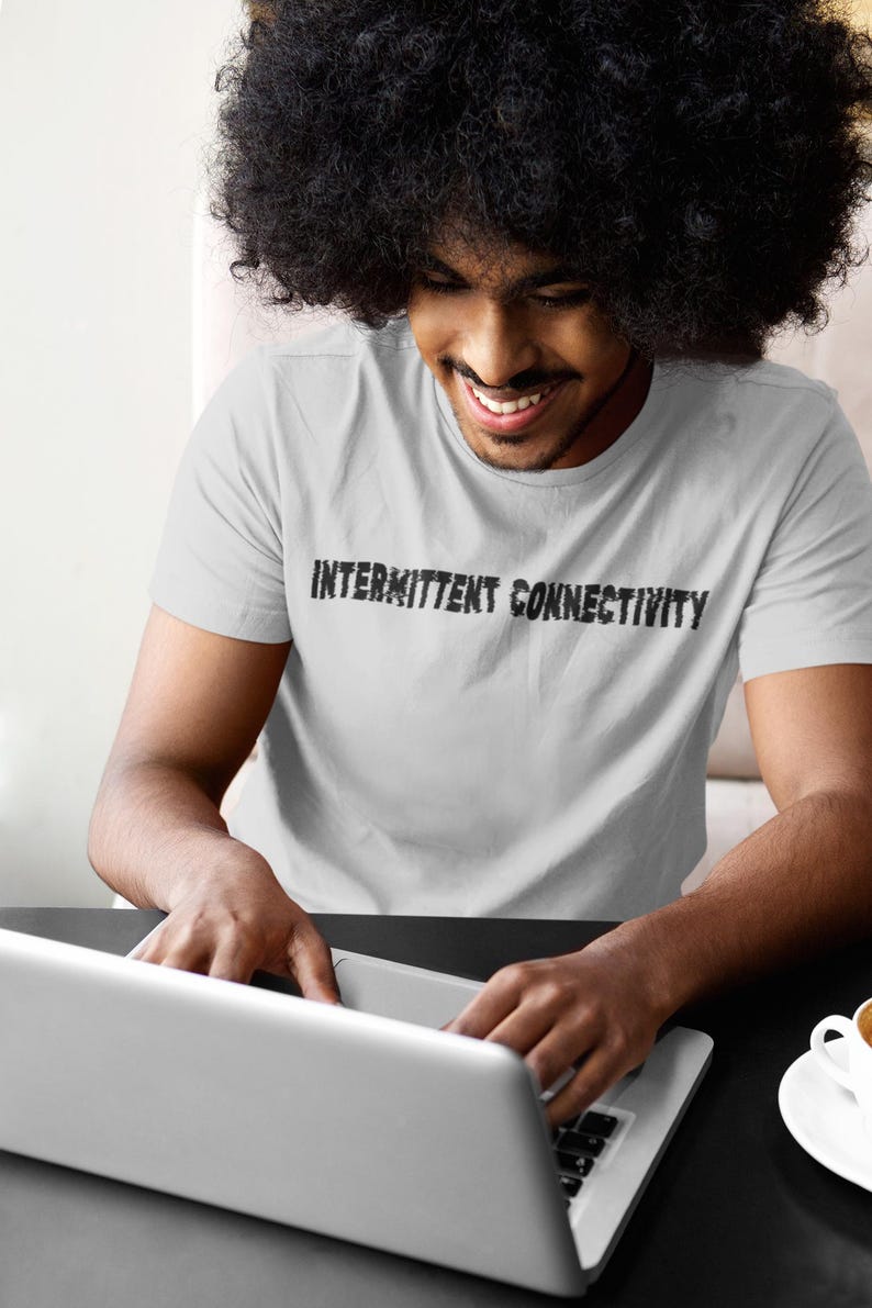 Puede incluir: Una persona que lleva una camiseta blanca con el texto "Intermittent Connectivity" impreso en ella, est&aacute; usando una computadora port&aacute;til. La persona est&aacute; sentada en una mesa con una taza de caf&eacute; sobre la mesa.