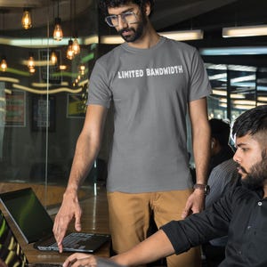 Pode incluir: Duas pessoas est&atilde;o trabalhando em laptops em um ambiente de escrit&oacute;rio. Uma pessoa est&aacute; vestindo uma camiseta cinza com o texto "Limited Bandwidth" impresso nela. A outra pessoa est&aacute; vestindo uma camisa preta de bot&otilde;es.