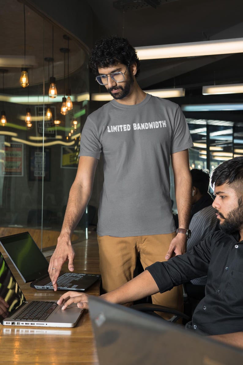 Pode incluir: Duas pessoas est&atilde;o trabalhando em laptops em um ambiente de escrit&oacute;rio. Uma pessoa est&aacute; vestindo uma camiseta cinza com o texto "Limited Bandwidth" impresso nela. A outra pessoa est&aacute; vestindo uma camisa preta de bot&otilde;es.
