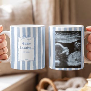 May include: Two white ceramic mugs with light blue vertical stripes. One mug features a rectangular frame with the text "Auntie Loading... EST. 2026" in blue. The other mug displays a black and white ultrasound image of a fetus inside a frame.