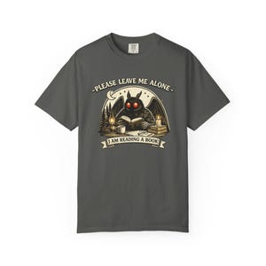 May include: Dark gray t-shirt featuring a cartoon illustration of a moth-like creature with red eyes reading a book. The text on the shirt reads "PLEASE LEAVE ME ALONE - I AM READING A BOOK."