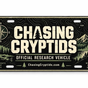 Puede incluir: Una placa de matrícula rectangular negra con el texto CHASING CRYPTIDS y OFFICIAL RESEARCH VEHICLE. El diseño incluye una rosa de los vientos, un paisaje montañoso, la silueta de una criatura, pinos y el sitio web ChasingCryptids.com.