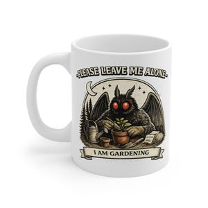 Puede incluir: Taza de cerámica blanca con una ilustración humorística de una criatura parecida a una polilla que está jardinería. La criatura tiene ojos rojos y alas grandes. El texto en la taza dice "PLEASE LEAVE ME ALONE" y "I AM GARDENING".