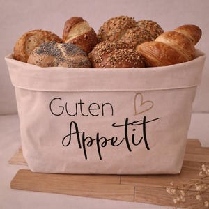 Könnte beinhalten: Ein Brotbeutel aus Leinen, gefüllt mit verschiedenen Backwaren, darunter Brötchen und Croissants. Auf dem Beutel steht in schwarzer Schrift "Guten Appetit" mit einem kleinen goldenen Herz. Der Beutel steht auf einer Holzoberfläche.