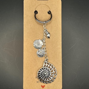 May include: A silver-coloured keyring with seashell charms. The keyring includes a large nautilus shell, a scallop shell, and a starfish charm, all suspended from a metal ring. The keyring is attached to a brown card with a heart symbol.