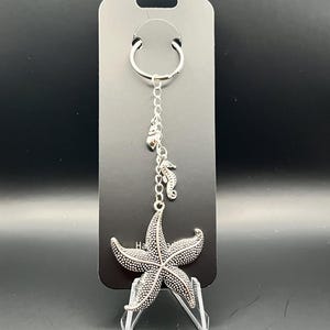 Può includere: Portachiavi a forma di stella marina argentata con un ciondolo a forma di cavalluccio marino e un piccolo ciondolo a cuore. Il portachiavi è attaccato a un cartoncino nero. La stella marina ha una superficie strutturata.