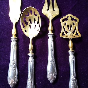 May include: Set of four antique serving utensils with ornate silver handles and gold-toned serving ends. Includes a butter knife, slotted spoon, fork, and cake server. The handles are engraved with floral patterns. The utensils are displayed on a purple velvet surface.