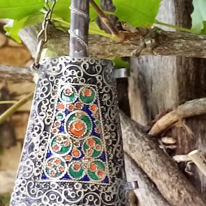 Puede incluir: Una campana de metal plateado con intrincados filigranas y un panel central decorado con vibrantes acentos de esmalte verde, naranja y azul. La campana tiene un mango superior cilíndrico y está suspendida por una fina cadena de metal.