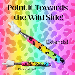 May include: Two colorful extendable pens with a peace sign on the top. The pens are decorated with a rainbow glitter leopard print. One pen is personalized with the text "Ms. Rubino". The text "Point it Towards the Wild Side!" and "Extends!" are visible in the image. The pens are on a rainbow leopard print background.