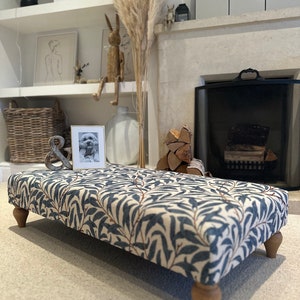 May include: A blue and white patterned footstool with wooden legs. The footstool is sitting on a light grey carpet in front of a fireplace.