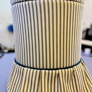 Könnte beinhalten: Ein zylindrischer Hocker mit einem gestreiften Muster in Creme und Dunkelgrün. Der Hocker hat einen plissierten Rock am Boden und einen passenden Deckel. Eine dunkelgrüne Zierleiste akzentuiert den Rock und die Oberseite des Sockels.