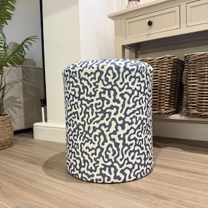 Könnte beinhalten: Ein zylindrischer Hocker mit einem marineblauen und cremefarbenen Korallenmuster. Der gepolsterte Hocker ist ein dekoratives Element. Der Hocker steht auf einem hellen Holzboden. Im Hintergrund sind ein Korb und eine Topfpflanze zu sehen.