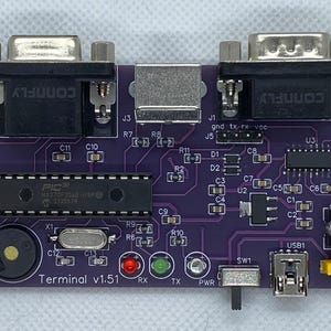 May include: A purple printed circuit board with a variety of components, including a PIC microcontroller, a buzzer, a USB port, a serial port, and a switch. The board is labeled "Terminal v1.51".