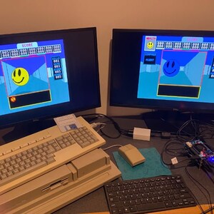 May include: Two computer monitors displaying a retro video game with a blue background and a yellow smiley face. The game has a score and health bar. A beige computer keyboard and a black keyboard are on a desk with a black and blue mouse pad.