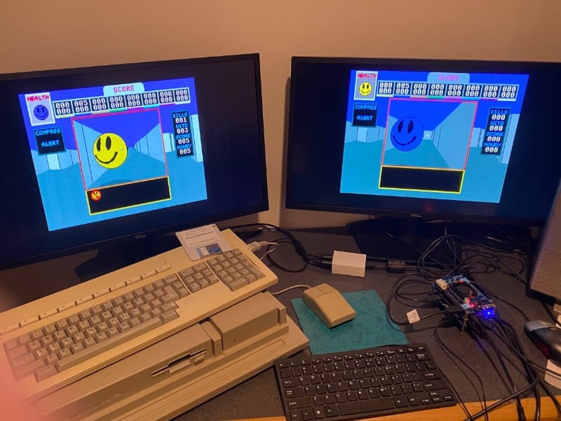 May include: Two computer monitors displaying a retro video game with a blue background and a yellow smiley face. The game has a score and health bar. A beige computer keyboard and a black keyboard are on a desk with a black and blue mouse pad.