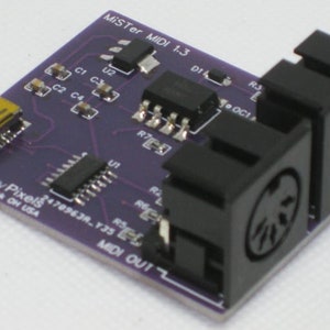 May include: A purple circuit board with the text "MiSTer MIDI 1.3" and "Legacy Pixels" printed on it. The board has two black MIDI connectors, a USB connector, and various electronic components.
