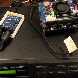 May include: A black Roland MT-32 Multi Timbre Sound Module with a green LCD screen displaying "MT-32" and "MIDI MESSAGE". The module has several buttons and knobs, including "PART", "SOUND GROUP", "VOLUME", "MASTER VOLUME", and "SELECT VOLUME". There is a blue circuit board with a fan and several USB ports, and a white box with two black audio jacks.