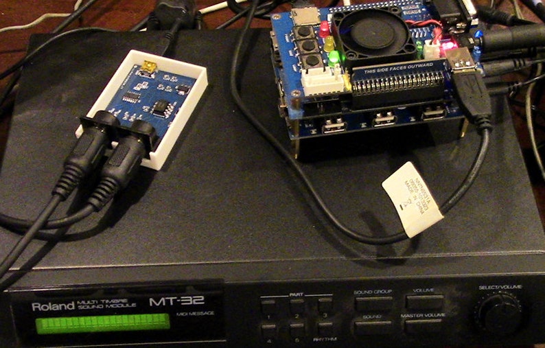 May include: A black Roland MT-32 Multi Timbre Sound Module with a green LCD screen displaying "MT-32" and "MIDI MESSAGE". The module has several buttons and knobs, including "PART", "SOUND GROUP", "VOLUME", "MASTER VOLUME", and "SELECT VOLUME". There is a blue circuit board with a fan and several USB ports, and a white box with two black audio jacks.