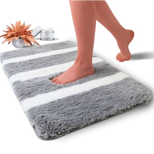 May include: A rectangular gray and white striped bath mat. The mat has a plush, textured surface with thick, soft fibers. A person's foot is stepping onto the mat. Decorative items are in the background.