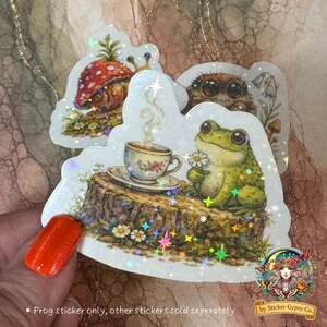 Close-up of holographic frog sticker held in hand, showing cottagecore frog with teacup and daisy, with other woodland stickers softly visible in the background.